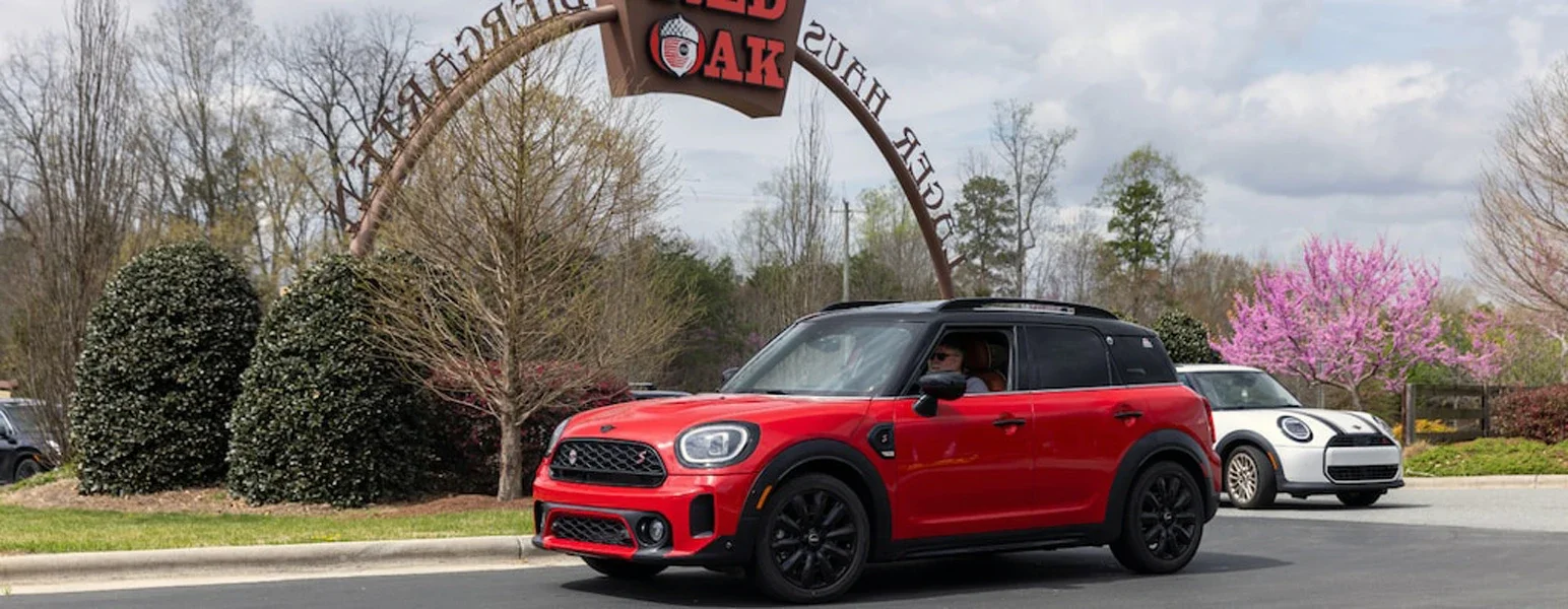 Red Mini Countryman driving past a 'RED OAK BREW HAUS' archway sign, with a white Mini parked behind it.