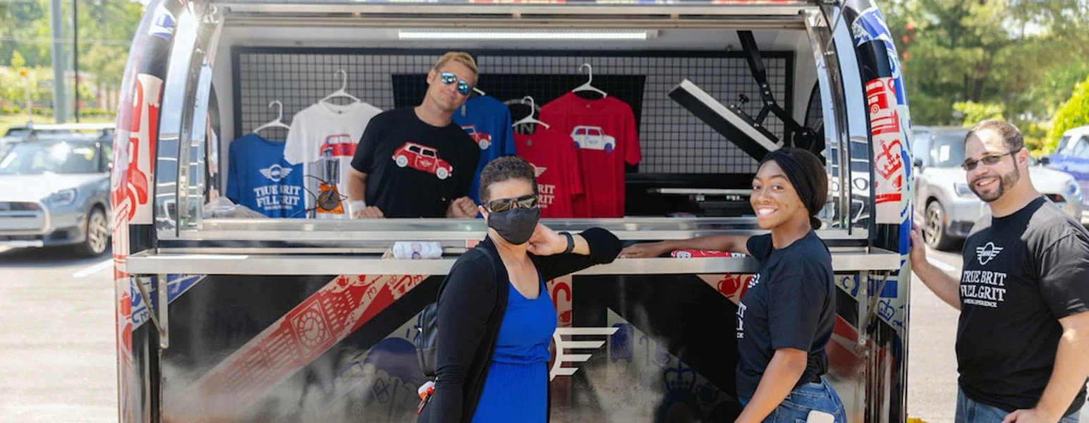 Four people stand around a mobile merchandise stand decorated with a Union Jack pattern, selling 'True Brit' shirts.