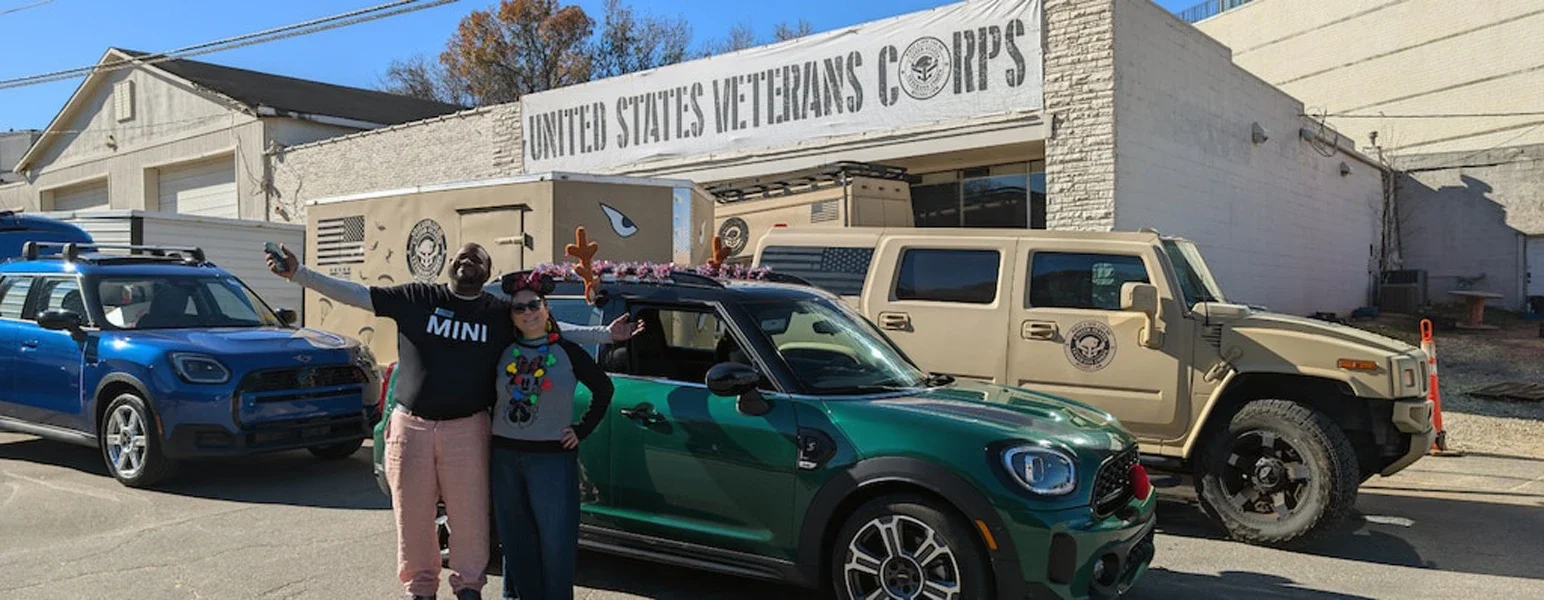 Two people pose with a decorated green Mini Cooper. In the background are a blue Mini, a tan Hummer, and a 'United States Vet'